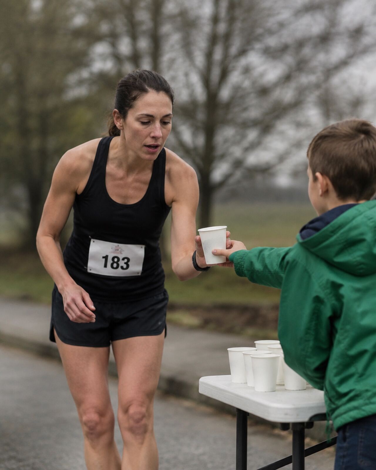 A young volunteer at a water station holding out a cup to a passing runner